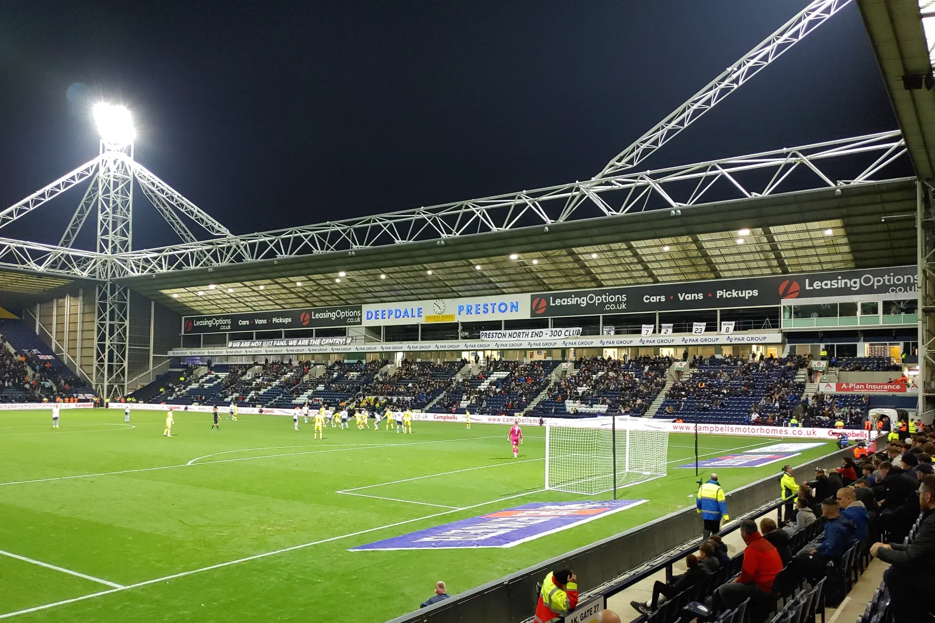 A floodlit football match at night, seen from the side of the pitch. Fans fill the stand opposite under a steel roof.