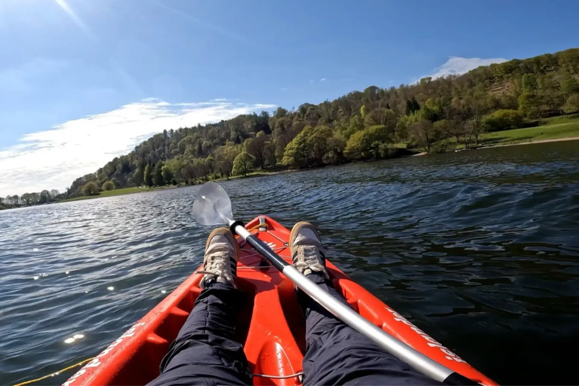 The front of a red kayak on a lake on a sunny day, with a paddle across it. Wooded hills rise from the far shore.